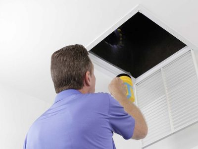 Older male with a yellow flashlight examining HVAC ducts in a large square vent. Male technician looking over the air ducts inside a home air intake vent.